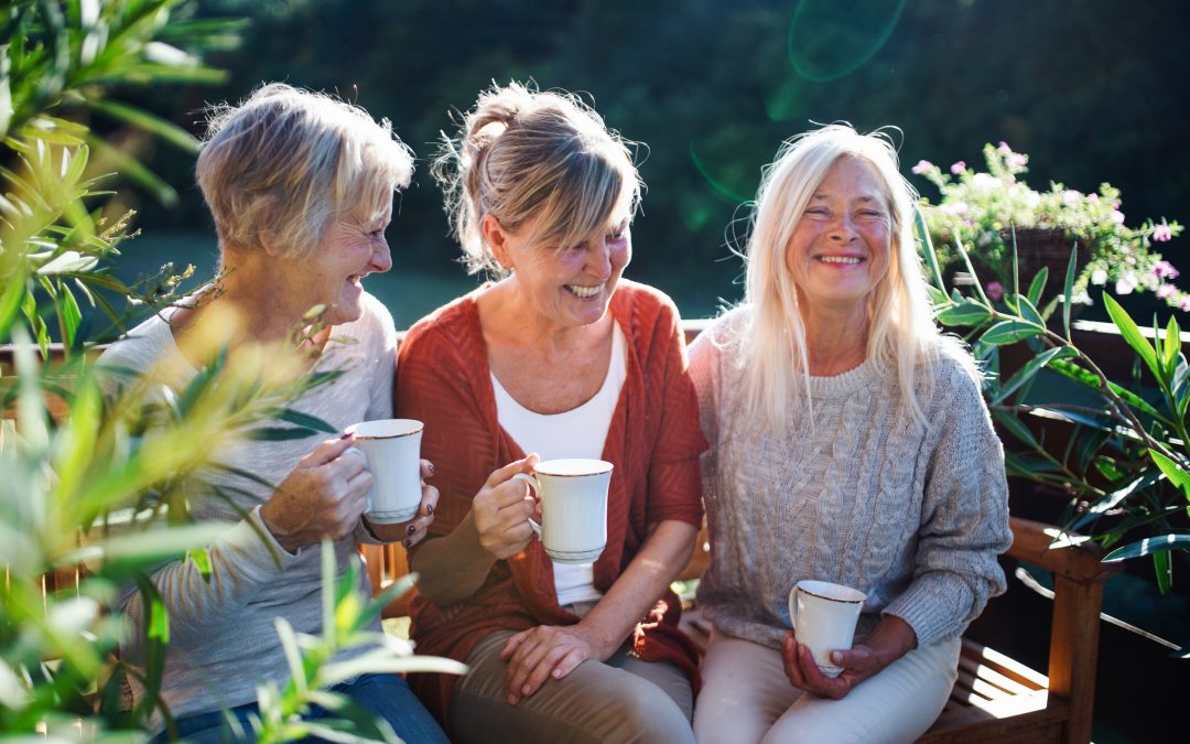 Three mature women gather in a sunlit garden, sharing laughter and warm cups of tea